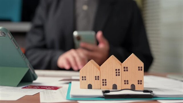 Three wooden house models sit in foreground businessman calculates mortgage payment options behind desk, holding mobile phone, reviewing financial documents, planning real estate investment.