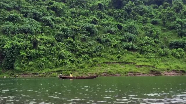 Video of wooden boat moving on river along dense forested hill a small with passengers cruising slowly a calm bordered by a lush green heavily hillside