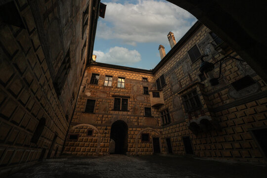 dramatic low-angle view of a historic castle courtyard featuring ornate Renaissance sgraffito facades, warm sunlight, and an arched gateway under a cloudy blue sky.