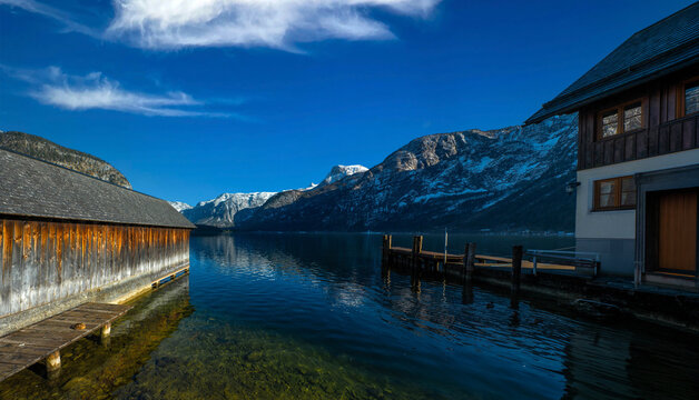 A serene view of Lake Hallstatt in Austria, framed by traditional wooden boathouses and a dock, with majestic snow-capped mountains reflecting in the clear water under a brilliant blue sky.
