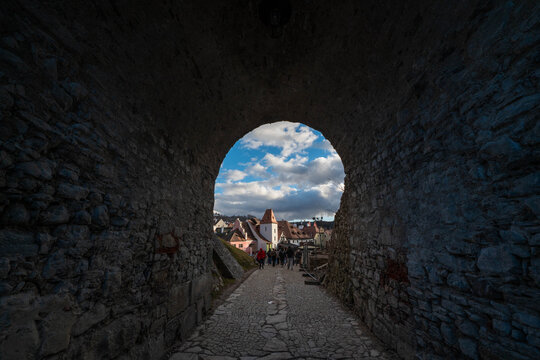 A dark historic stone archway framing a view of a charming old town street with traditional buildings, cobblestone paths, and people walking under a bright cloudy blue sky