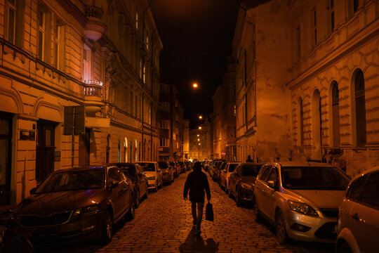 A lone man walking down a historic cobblestone street lined with parked cars and glowing old buildings at night urban atmosphere.