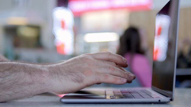 Hand types on laptop keyboard in public space. Background shows blurred cafe patrons and glowing signs. Focus stays on fingers and laptop. Person appears focused and relaxed while working