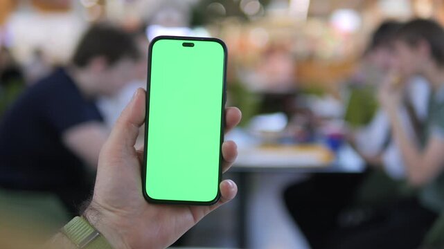 Hand holds smartphone with green screen. Background shows blurred cafe scene. People sit at tables, eating or talking casually. Atmosphere feels relaxed and social. Perfect for apps, marketing