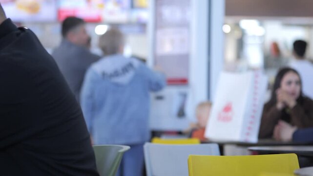 Blurry indoor scene with people sitting and standing near tables. One person points at a menu board. Others chat or wait. Yellow chairs add color to space. Suitable for cafe, restaurant
