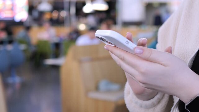Hands hold phone in cafe setting. Person focused on screen, relaxed posture. Background shows blurred dining area. Ideal for social media, marketing, or lifestyle content