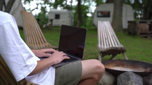 Person typing on laptop while seated in a hammock. Background shows campsite with trailers and firepit. Person appears relaxed, focused on work or leisure. Ideal for remote work, travel