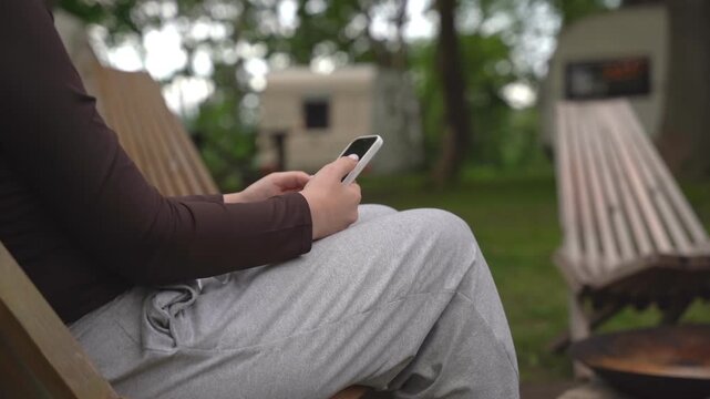 Person relaxes on wooden chair, using smartphone outdoors. They sit calmly, focused on phone screen. Background shows grassy yard and campfire pit. Ideal for scenes of leisure, digital connection
