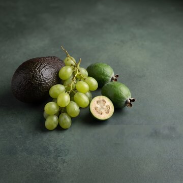 Ripe avocado, fresh green grapes, and feijoa fruits arranged naturally on a muted dark green background, ultra realistic studio food photography, premium commercial style