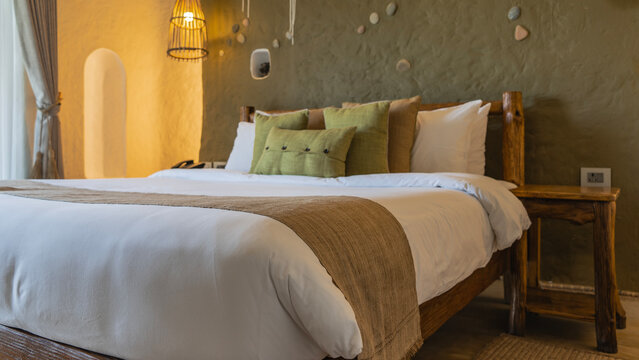The bedroom interior is rustic. The bed is made up. Pillows at the headboard. Log bedside tables. The wall is roughly finished and decorated with pebbles. The lamp is lit.
