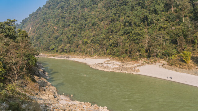 Mountain river. A green forest grows on the coastal hills. Silhouettes of people walking are visible on the sandy shore. The blue sky. Nepal. Seti River   