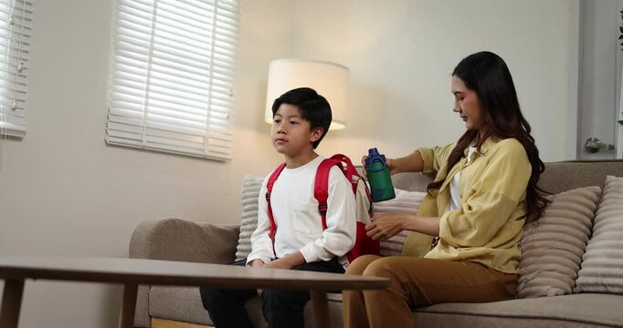 Asian mother and son preparing for school in a bright home setting, with the parent helping the child pack his red backpack and water bottle, showcasing family support and education responsibilities
