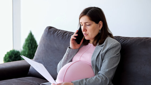 Pregnant businesswoman wearing a blazer and pink maternity dress, sitting on a sofa at home, talking on a smartphone while holding and reviewing documents