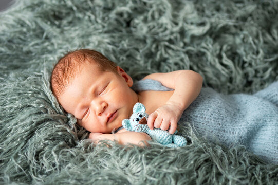 Newborn baby boy sleeping wrapped in a blue blanket on a soft green furry rug, holding a small knitted blue mouse toy, portraying innocence and early childhood dreams