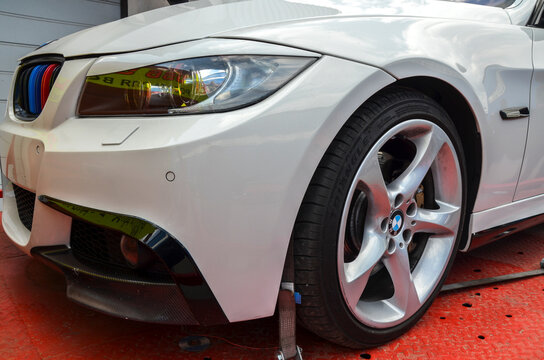 Close up of white BMW 3-Series E90 sedan front end. It highlights the sharp headlight design, five spoke alloy wheels, M-colored grille, and custom front lip spoiler