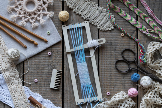 Rustic weaving and macrame craft scene with loom blue threads crochet hooks comb scissors lace and handmade textile accessories on wooden table flat lay