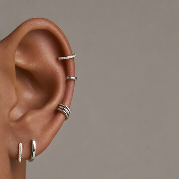 Close-up of human ear with stylish silver piercing jewelry, cuffs, and studs on a minimalist solid background