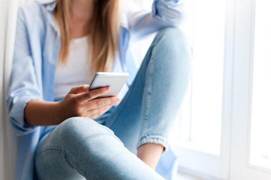 Young woman using smartphone at home, sitting by window. Girl browsing, shopping. Distance learning, online education, connection, communication. Doomscrolling, mobile addiction. Close up of hands