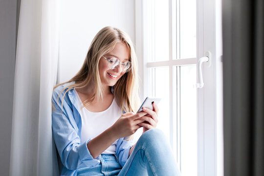 Young woman using smartphone, sitting by window. Relaxation on windowsill. Concept of shopping online, distance learning, banking. Freelancer working from home office. People connection, communication