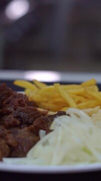 Slow motion camera panning of traditional roasted beef meat served with crispy french fries and sliced raw onions on a white plate, vertical video.