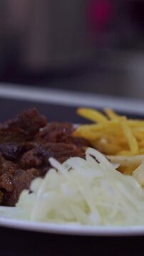 Slow motion camera panning of traditional roasted beef meat served with crispy french fries and sliced raw onions on a white plate, vertical video.