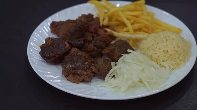 Slow motion camera panning of traditional kazan kebab with roasted meat, crispy french fries, rice side dish, and sliced raw onions on a white plate, horizontal video.