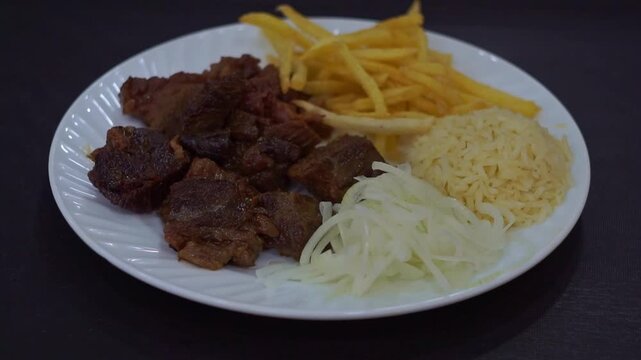 Slow motion camera panning of traditional kazan kebab with roasted meat, crispy french fries, rice side dish, and sliced raw onions on a white plate, horizontal video.