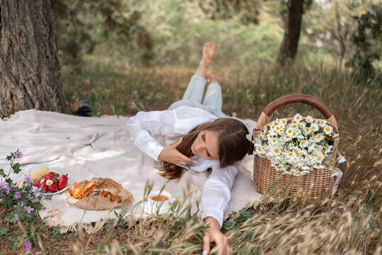 Young woman resting on a picnic blanket with fresh strawberries and flowers in a park during a sunny afternoon