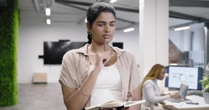 Early-adult Asian Indian woman opening planner and writing notes while planning at open-plan office
