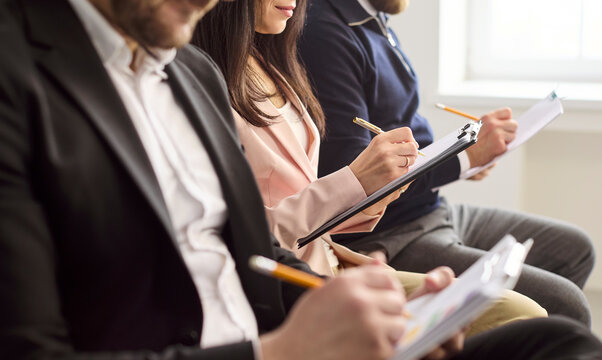 Candidates in interview recruitment hiring assessment waiting. Business applicants write notes on clipboards and forms in a bright office. Clear concept of professional hiring process.