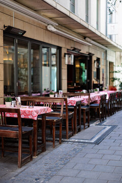 Empty wooden tables and chairs with pink checked cloths set out along a narrow city street outside a restaurant on a summer morning.