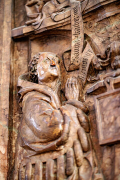 Carved wooden relief of a tonsured monk gazing upward while clutching a cross inscribed with Gothic lettering.
