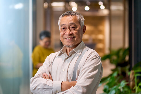 Happy Senior Asian Man with Suspenders Smiling with Arms Crossed in Modern Office, Confident Elderly Male Professional, Active Retirement Portrait