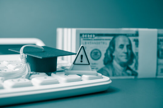 Warning sign and mortarboard on white calculator and heap US dollar banknotes. Student loan or student debt non payment, delinquent, bad credit score, financial crisis, education concept.