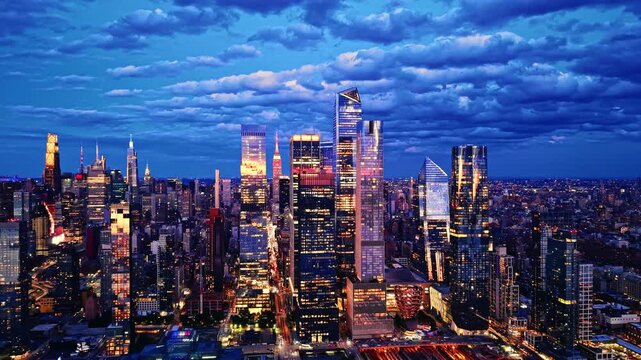 Hudson Yards skyline beneath cloudy dusk sky, New York City. Glass towers reflect city lights under dramatic clouds at evening hour. Aerial view.