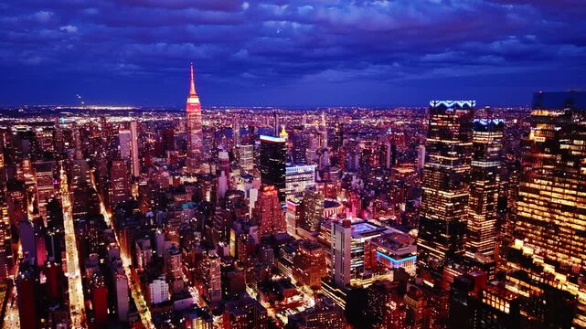 Night skyline with building spire lit red, Manhattan. Storm clouds gather above illuminated towers and city streets below. Aerial view.