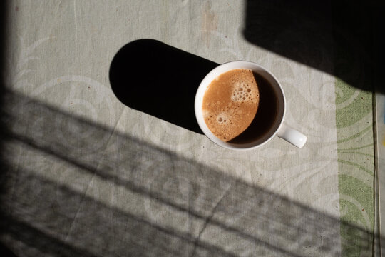 A cup of morning coffee with foam and a long shadow in warm sunlight. Still life.
