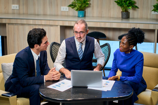 Diverse Team of Business Professionals Collaborating in Modern Office, Senior Executive Mentoring Colleagues During Meeting with Laptop and Charts