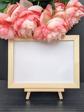 Charming wooden frame on an easel surrounded by vibrant pink peonies, creating a beautiful and artistic display, perfect for showcasing messages, photographs, or art pieces in a cozy, elegant set