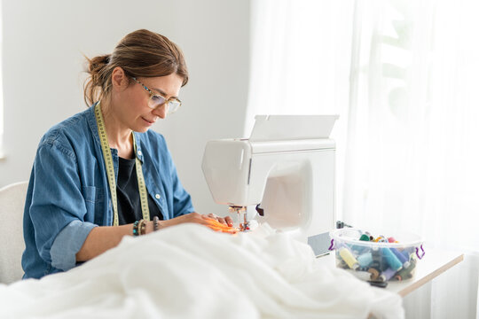 Woman sews fabric on white machine in bright daylight.