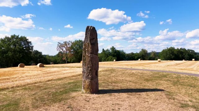 Pr&auml;historischer Gollenstein-Menhir auf einer Wiese bei sommerlicher Erntezeit mit Strohballen unter blauem Wolkenhimmel. Altes Kulturdenkmal in Mitteleuropa.