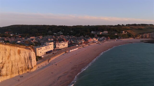 The drone captures the seafront promenade and colorful beach huts at sunset in Etretat.