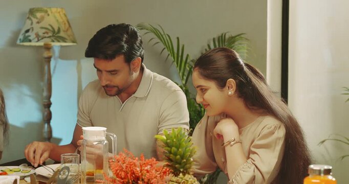 Indian happy man woman couple sitting around table chair talk speak share gossip room place young adult gen z male hold roti bowl serve fresh salad female eat tasty food spend time enjoy dinner night