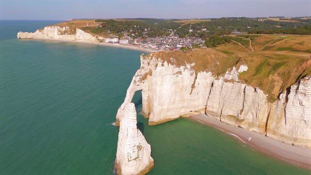 A drone pulls back to reveal a limestone arch and towering rock spire near Etretat, France.