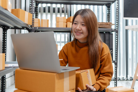 Young entrepreneur manages online orders with a laptop while checking shipping boxes in a warehouse, representing ecommerce, inventory management, order fulfillment, small business, and logistics.