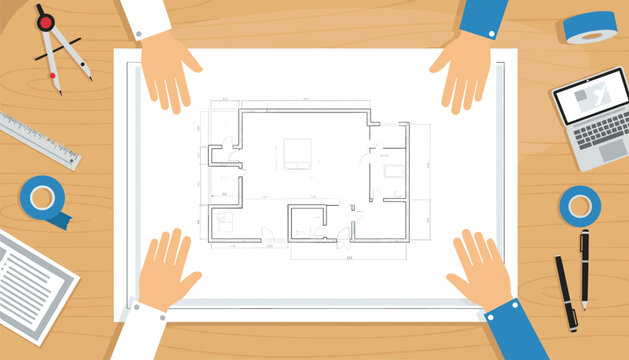 Top down view of architects working on a house floor plan blueprint on a wooden table with drafting tools, tape, and a laptop computer.