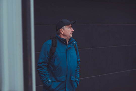 Thoughtful senior man aged 50s 60s in blue jacket and cap standing alone outdoors looking into distance, psychological mental health concept of loneliness and reflection