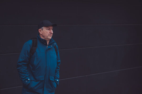 Thoughtful senior man aged 50s 60s in blue jacket and cap standing alone outdoors looking into distance, psychological mental health concept of loneliness and reflection