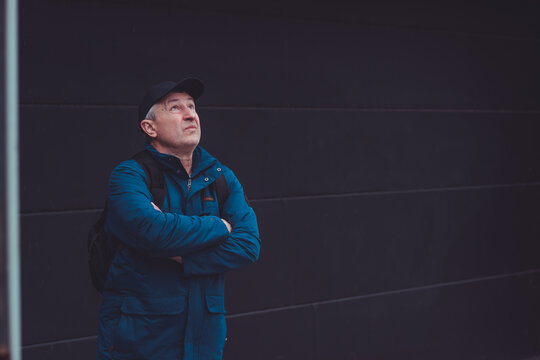Thoughtful senior man aged 50s 60s in blue jacket and cap standing alone outdoors looking into distance, psychological mental health concept of loneliness and reflection
