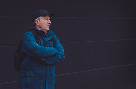 Thoughtful senior man aged 50s 60s in blue jacket and cap standing alone outdoors looking into distance, psychological mental health concept of loneliness and reflection